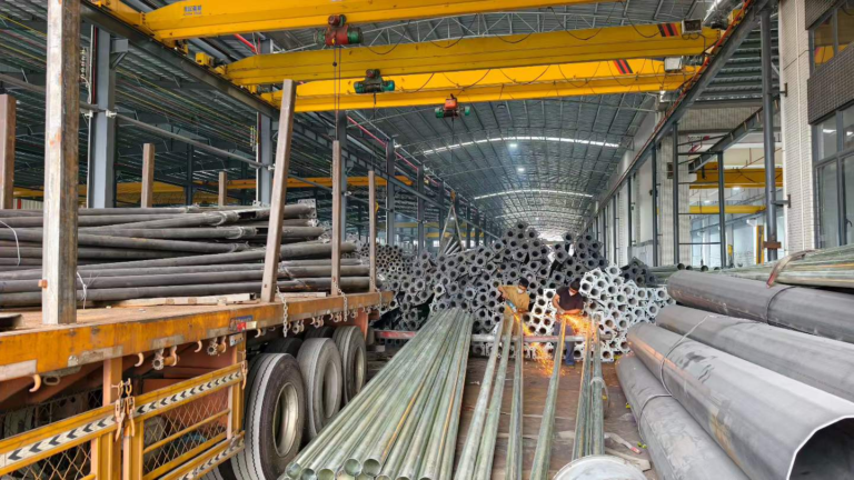 Workers grinding steel lighting poles beside stacked poles and a loaded truck inside a factory