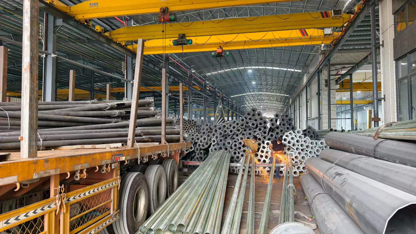 Workers grinding steel lighting poles beside stacked poles and a loaded truck inside a factory